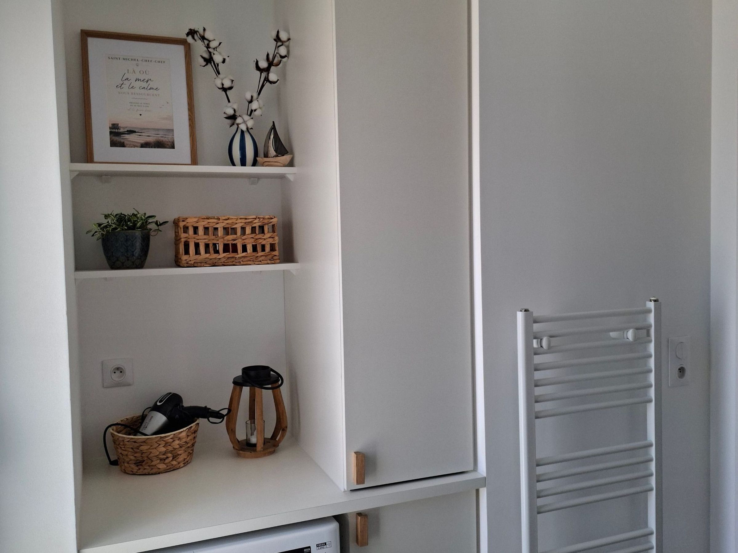 A white interior with a shelf holding a framed picture, a vase with branches, and a wicker basket with a lantern.