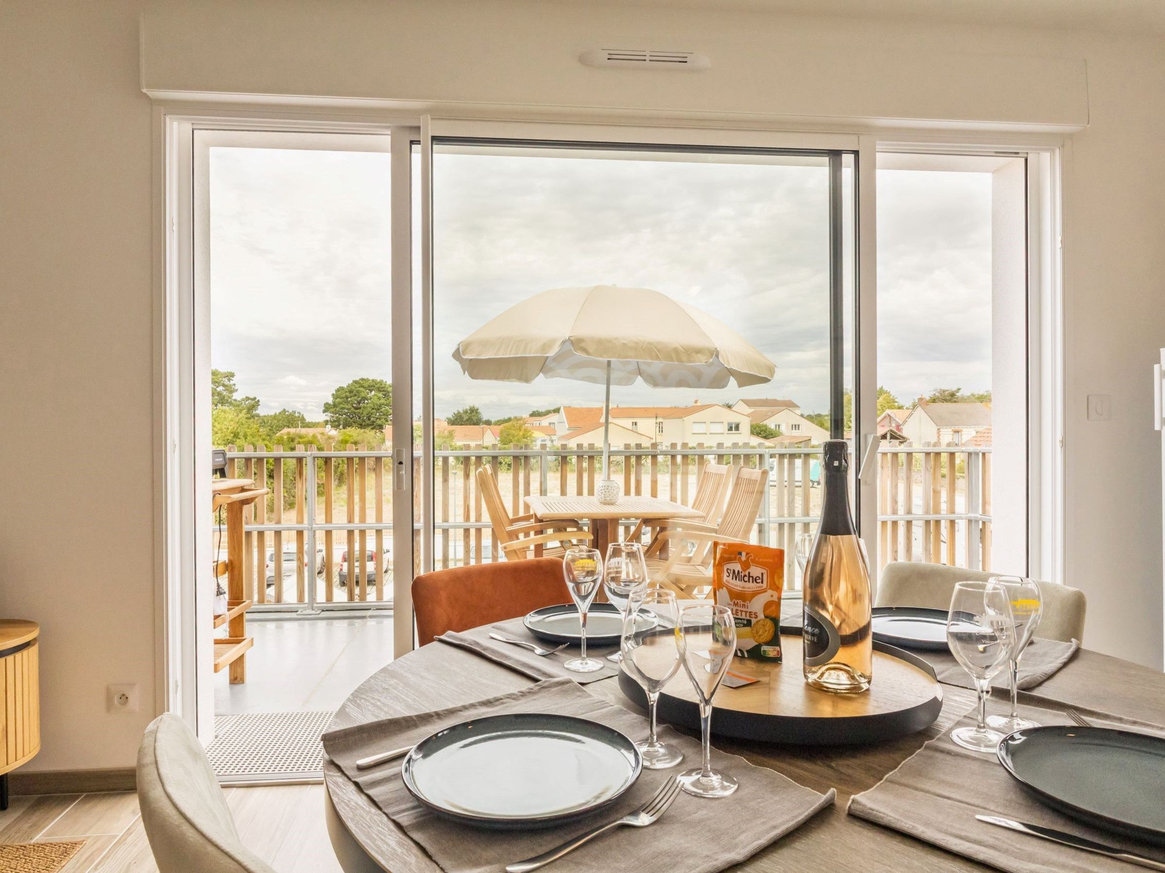 A dining area with a table set for four, a bottle of champagne, and a view of a balcony with a wooden fence and houses in the background.