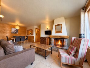 A cozy living room with a fireplace, a sofa, a striped armchair, and a dining area with a table and chairs. - Ski-in/ski-out apartment - 6 guests (Belle Plagne)