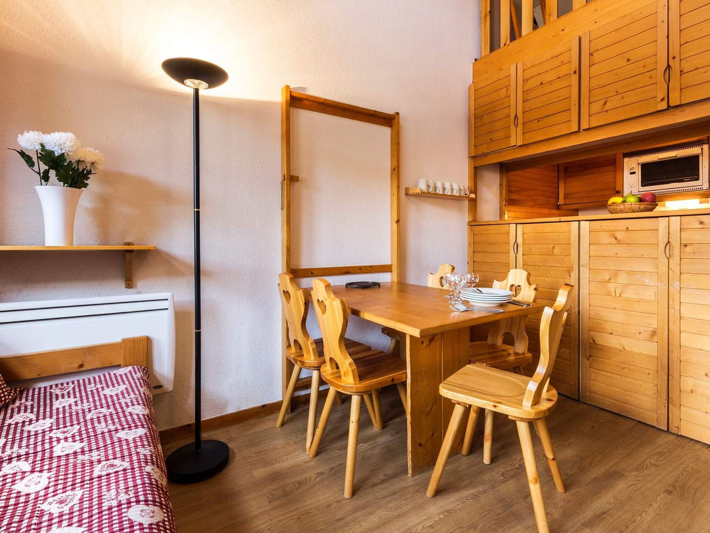 Una habitación compacta con una mesa de comedor de madera, cuatro sillas, una pequeña cocina con microondas y una cama individual con manta estampada.