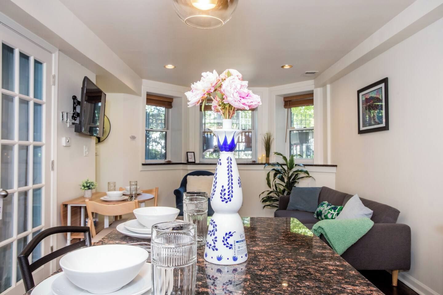 A dining area with a table set for two, a vase with flowers, and a television mounted on the wall.