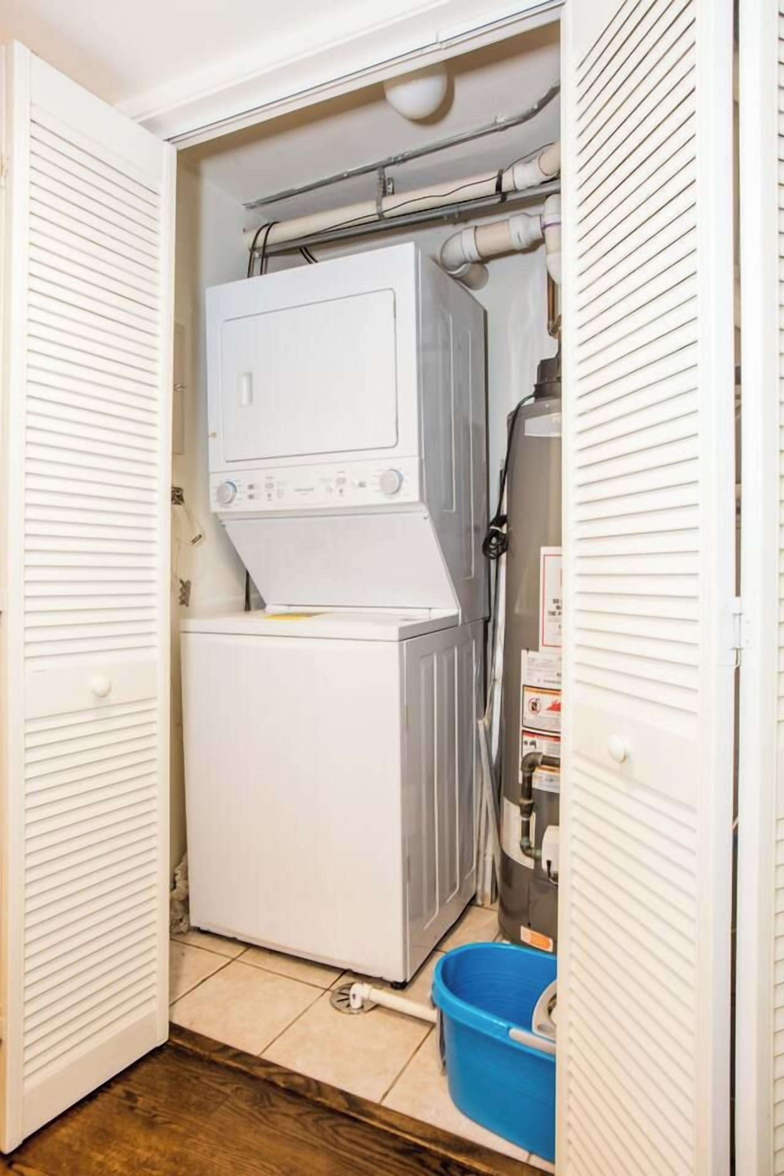 Une buanderie équipée d’un lave-linge et d’un sèche-linge superposés, d’un seau bleu et de tuyauteries apparentes.