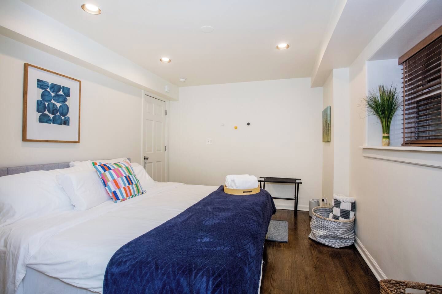 Une chambre d’hôtel avec un lit, une table de chevet, une fenêtre avec des stores, une œuvre d’art accrochée au mur et une petite plante posée sur le rebord de la fenêtre.