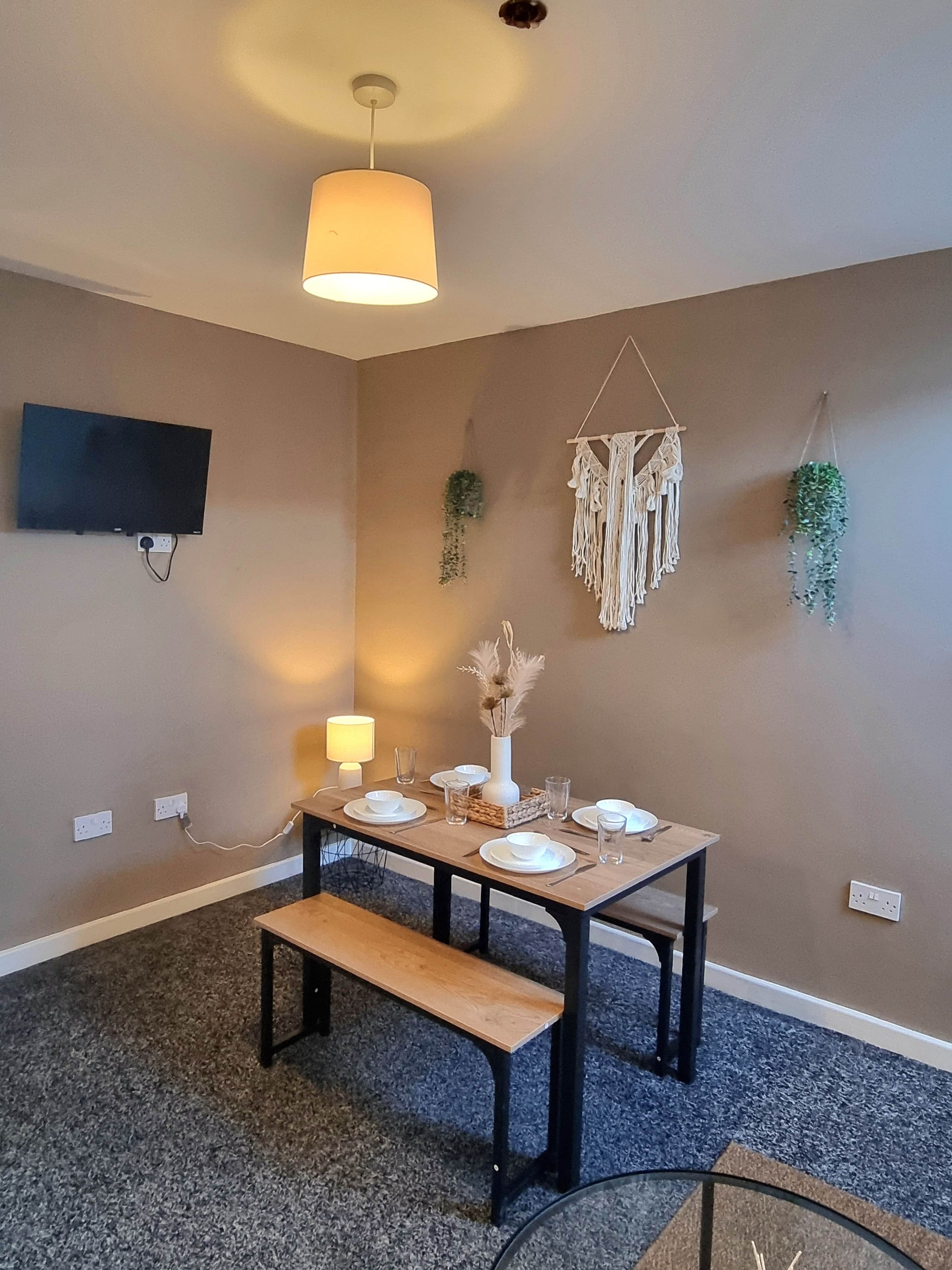 Een eethoek met een tafel voor vier personen, een bank, een aan de muur bevestigde televisie en een wanddecoratie van macramé.