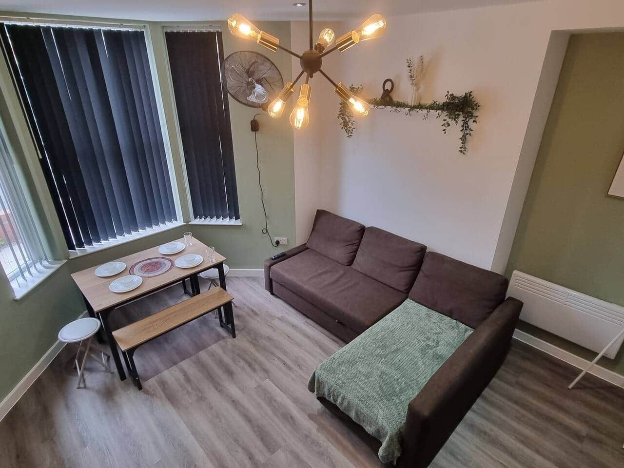 Una sala de estar con sofá, una mesa de comedor preparada para cuatro personas, un banco, un aplique de pared y un radiador.