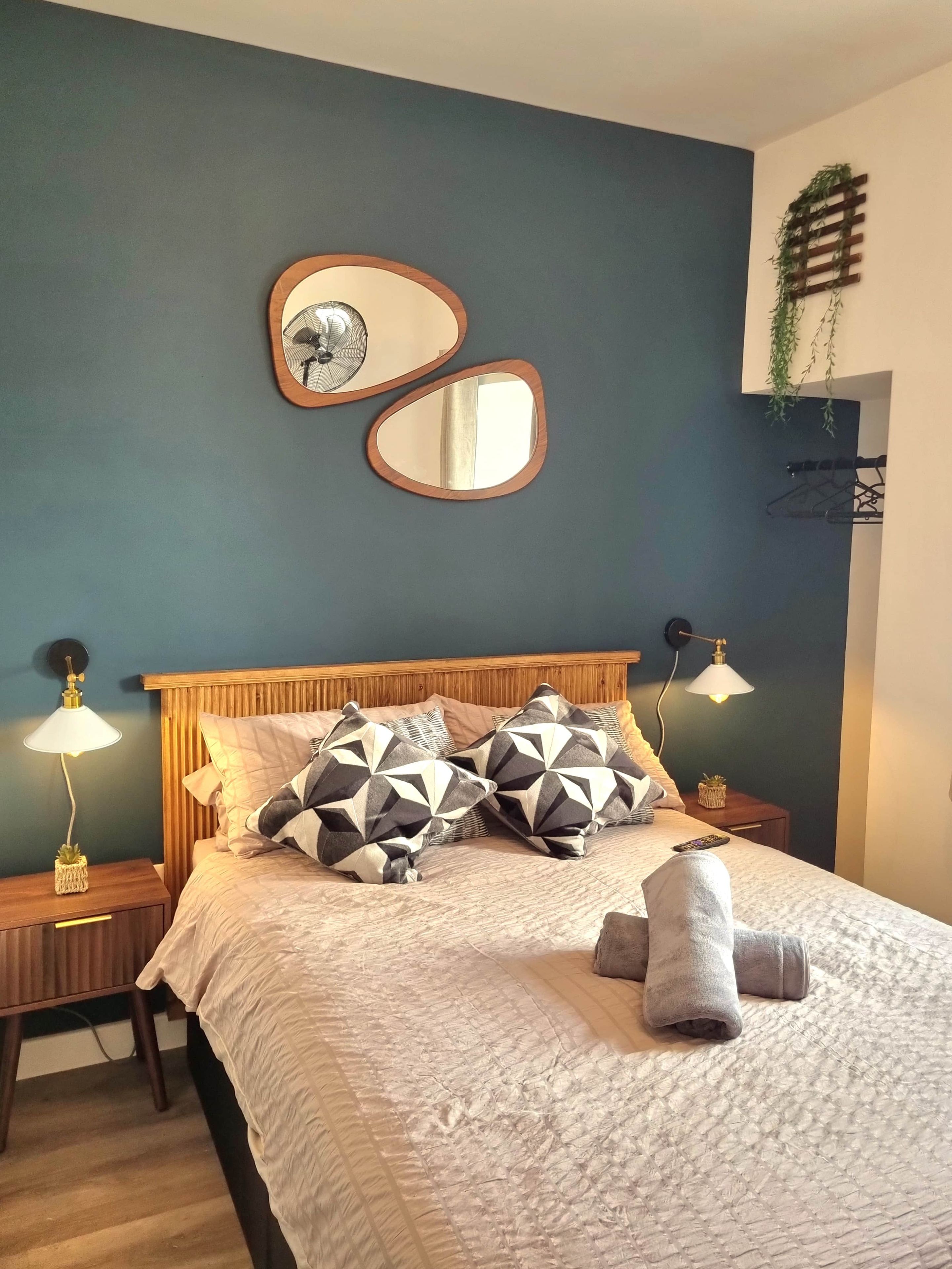 Une chambre à coucher avec un lit, deux tables de chevet, un miroir fixé au mur et une plante suspendue.