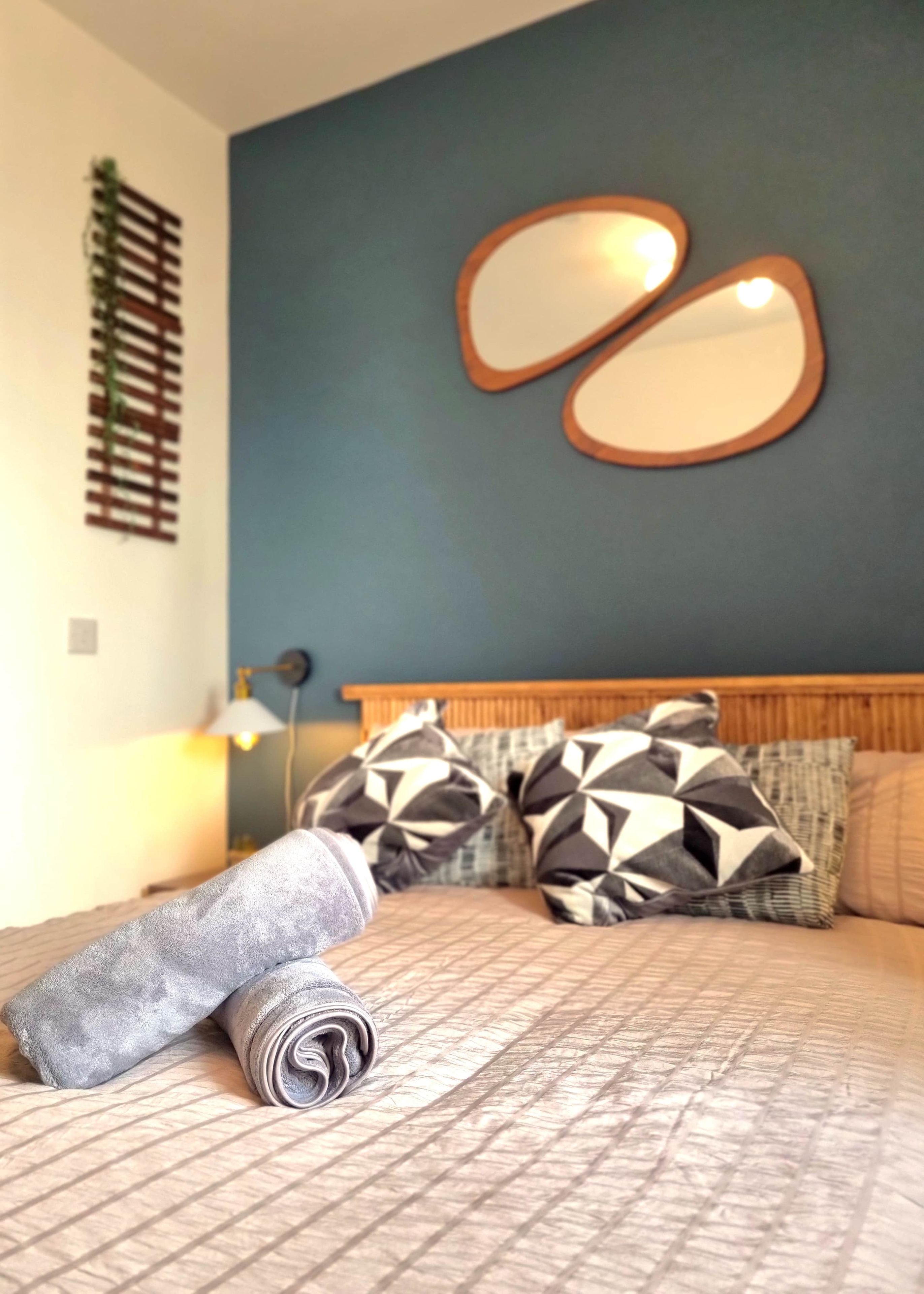 Una cama bien hecha con almohadas estampadas, un espejo en la pared y una lámpara de noche.