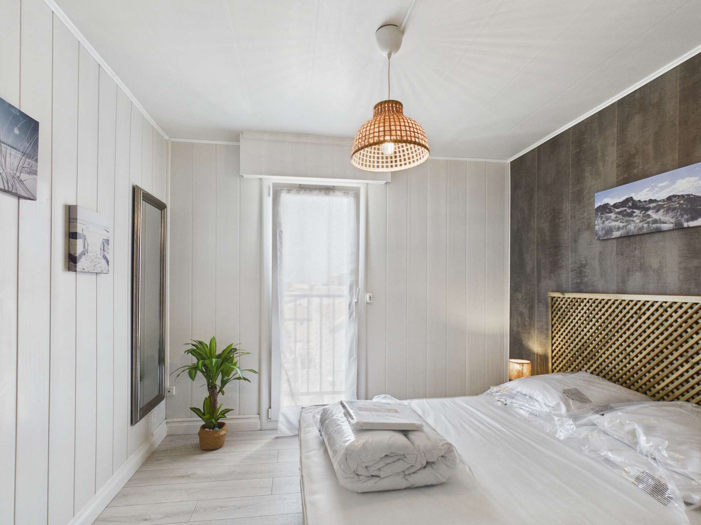 A bedroom with a bed, a potted plant, a hanging light fixture, and a door leading to another room.