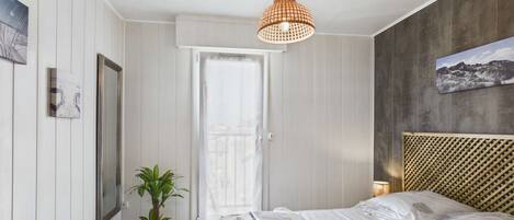 A bedroom with a bed, a potted plant, a hanging light fixture, and a door leading to another room.