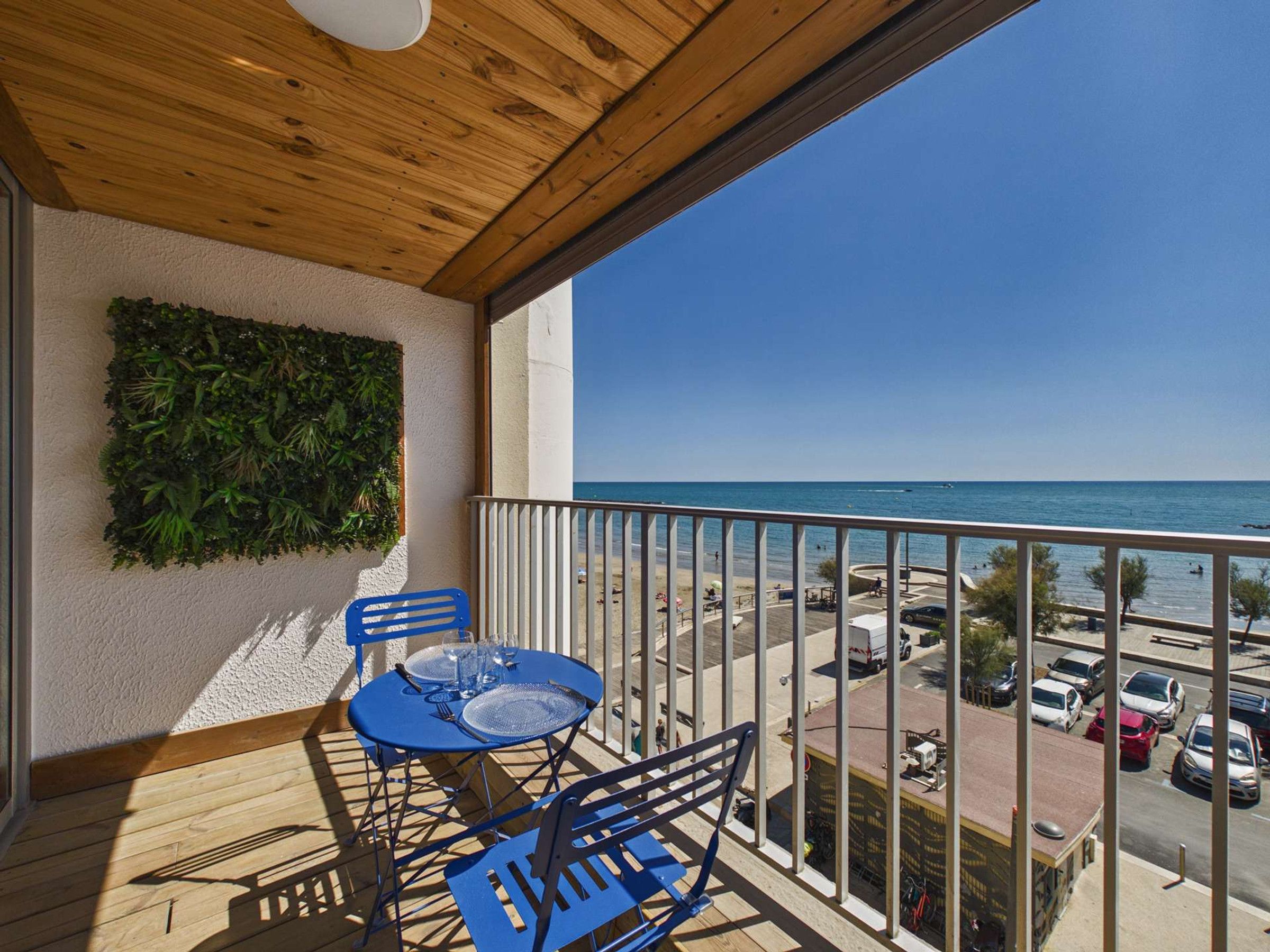 Ein Balkon mit Holzboden, ein blauer Tisch für zwei Personen und Blick auf das Meer und einen Parkplatz.