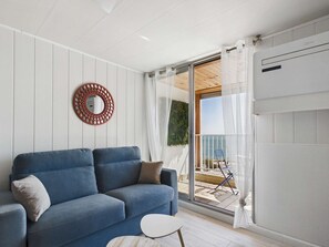 Una sala de estar moderna con un sofá azul, una mesita redonda y un ventanal que da a un área exterior con vista al mar.