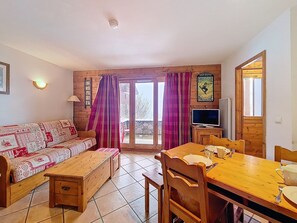 A cozy room with a wooden sofa, a wooden dining table with chairs, a television, and a window with curtains. - Les Menuires - 3 rooms, 6 pers, terrace, pool access (Les Menuires)