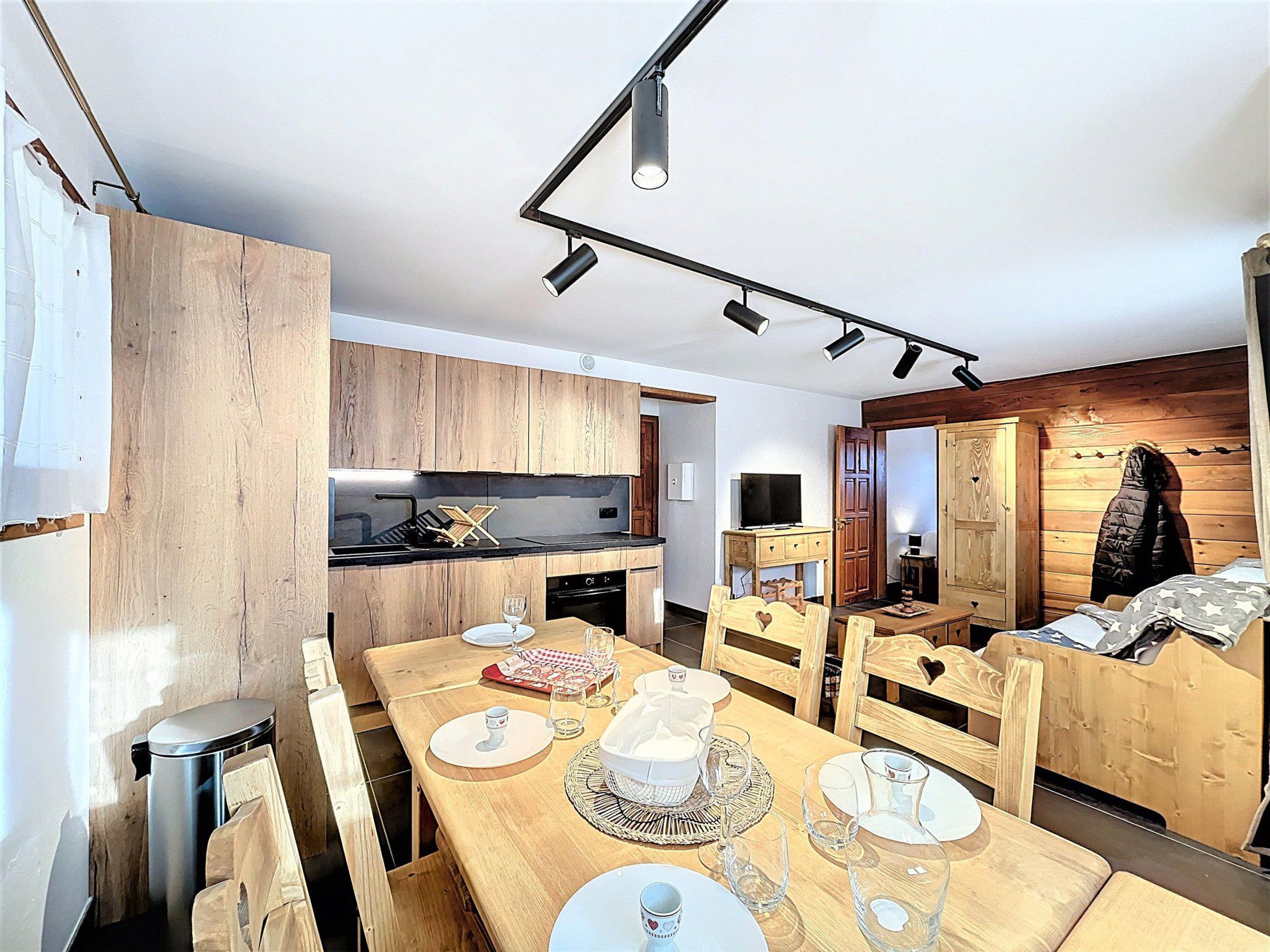 Une cuisine compacte avec des meubles en bois, un coin repas avec une table en bois pour quatre personnes, et un coin salon confortable avec un canapé et un porte-manteau.