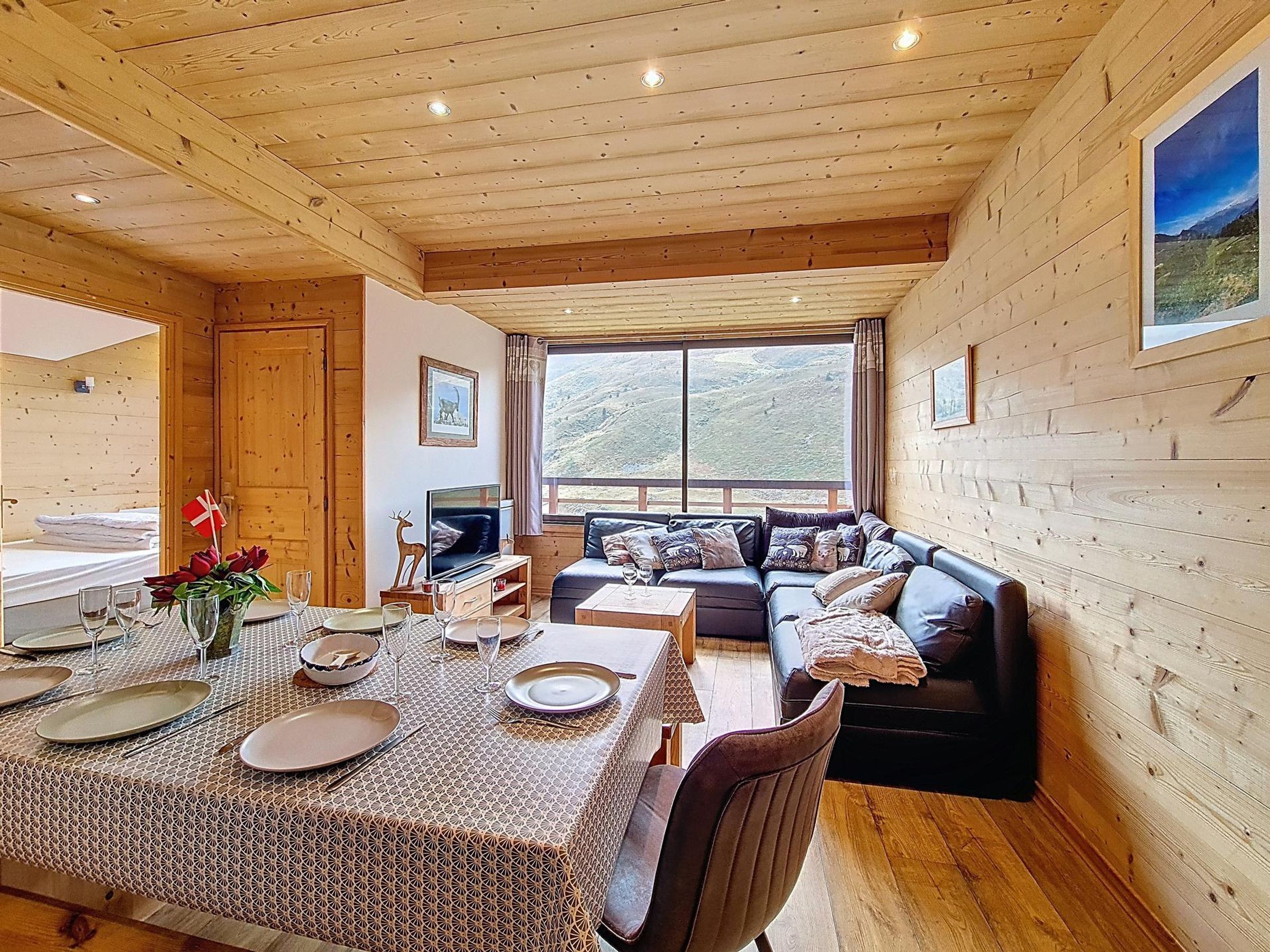 A wooden cabin interior with a dining area, sofa, and a large window offering a view of the outdoors.