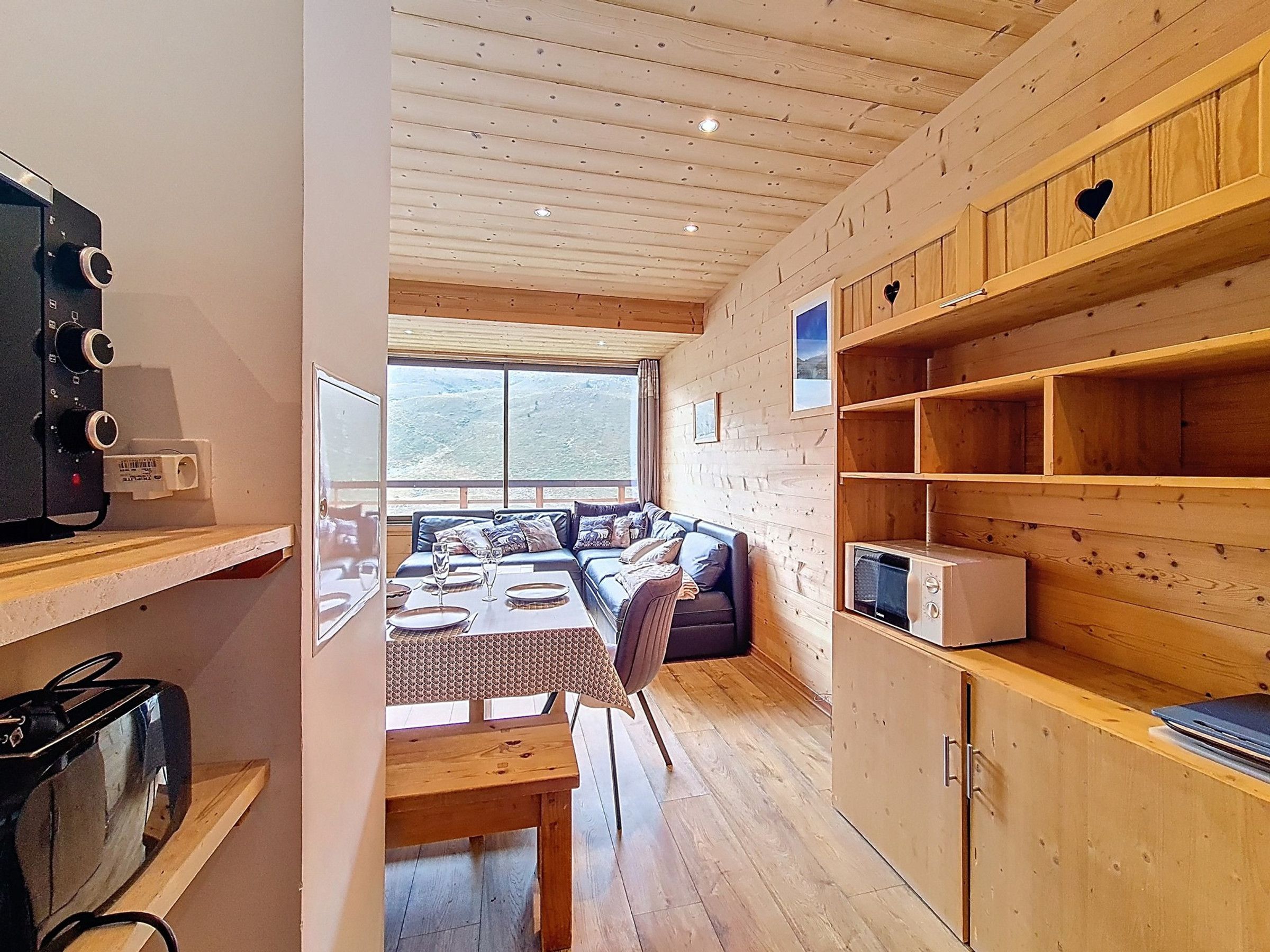 A cozy cabin interior with a wooden ceiling, a sofa with cushions, a dining table set for two, and a kitchenette area.