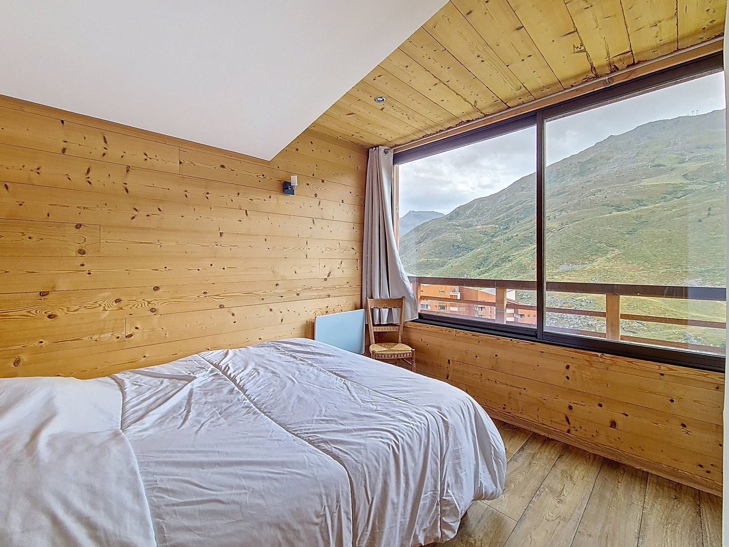 A bedroom with a large window, wooden walls, a bed, and a chair.