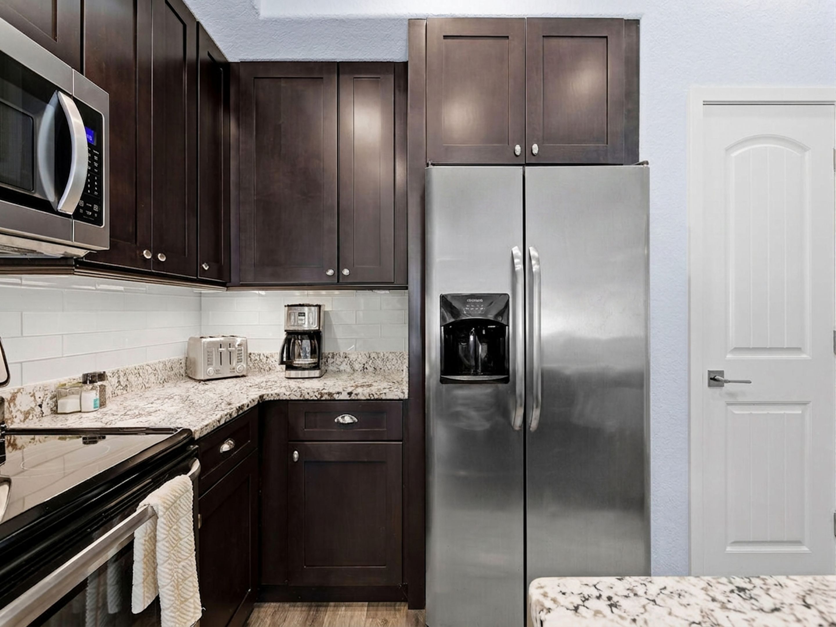 A compact kitchen with dark wood cabinets, a stainless steel refrigerator, a microwave, a toaster, and a coffee maker.