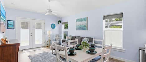 A dining area with a white table, blue cushioned chairs, a ceiling fan, and a view of the outdoors through a window.