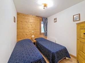 A small room with two single beds, a wooden wall, a window with curtains, and a wooden cabinet. - Spacious 3-room apartment near slopes with terrace, pool and parking (Les Menuires)