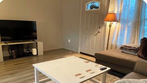 A living room with a flat-screen TV on a stand, a white coffee table, a brown sofa, and a lamp. - Cozy Home Away From Home (Woodbridge)