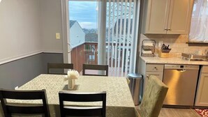 A dining area with a table set for four, a kitchen with a dishwasher, and a view through a sliding door. - Cozy Home Away From Home (Woodbridge)