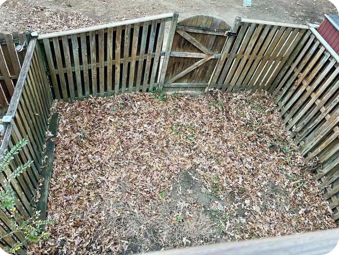 Ein von einem Zaun umgebenes Gebiet mit einem hölzernen Tor, bedeckt mit trockenem Laub und etwas Grün.