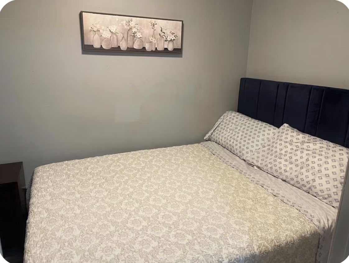 A bed with a patterned bedspread, a headboard, and a framed artwork on the wall.
