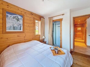 Ein Zimmer mit einem großen Bett, einem gerahmten Bild einer Berglandschaft, einem Fenster mit Vorhängen und einer Tür zu einem weiteren Raum.