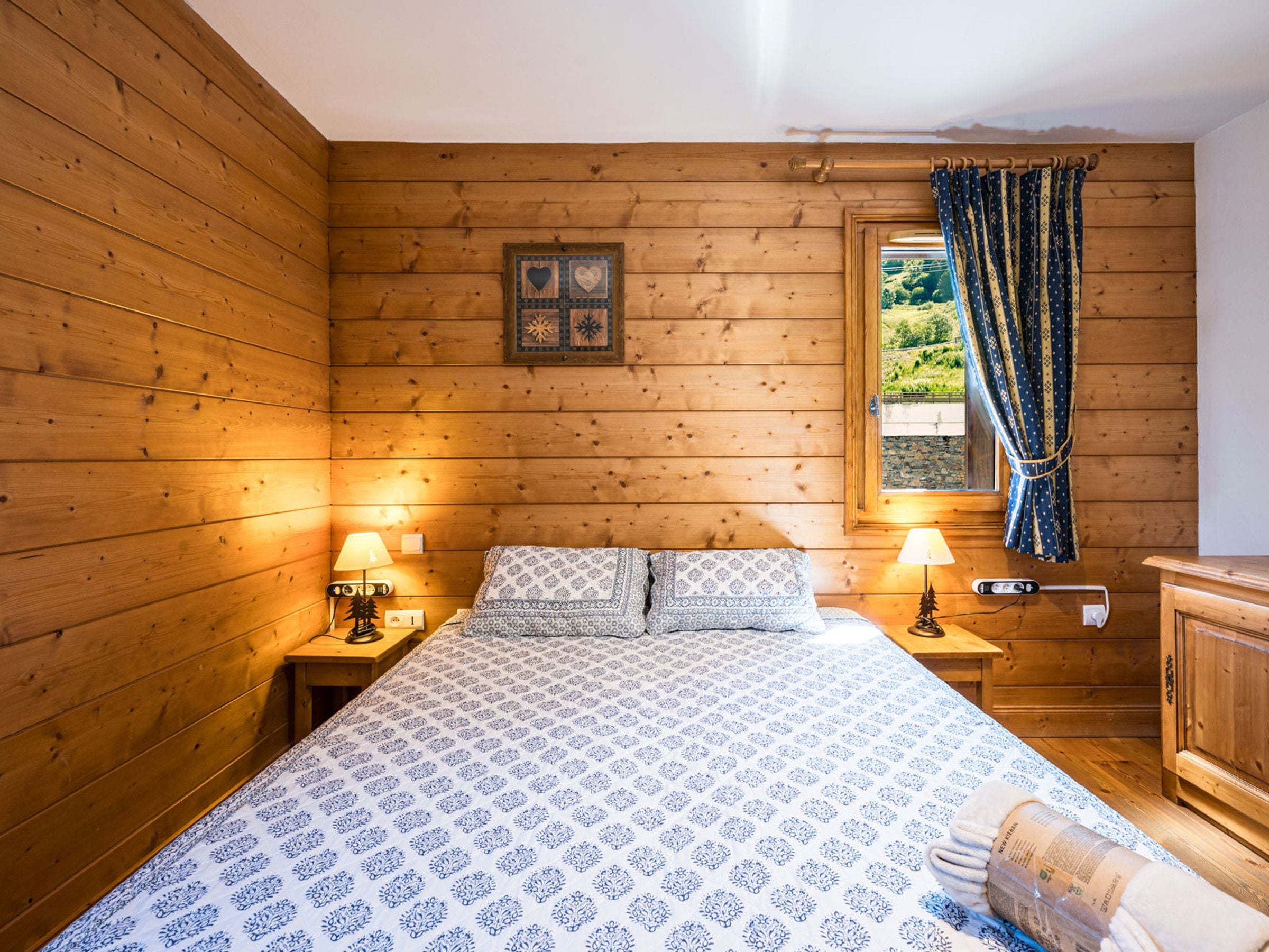 A wooden-paneled room with a bed, two bedside lamps, a window with curtains, and a wooden cabinet.