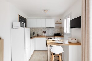 Eine kompakte Küche mit weißen Schränken, einer Mikrowelle, einem Toaster und einer Kaffeemaschine. Es gibt einen Essbereich mit einem weißen Stuhl und einem Tisch für zwei Personen.