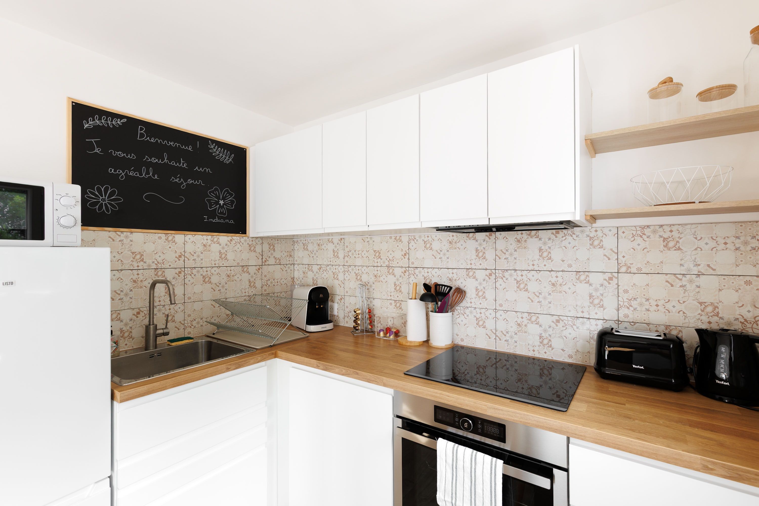 A modern kitchen with white cabinets, a blackboard with a welcome message, a coffee maker, a toaster, and a dishwasher.