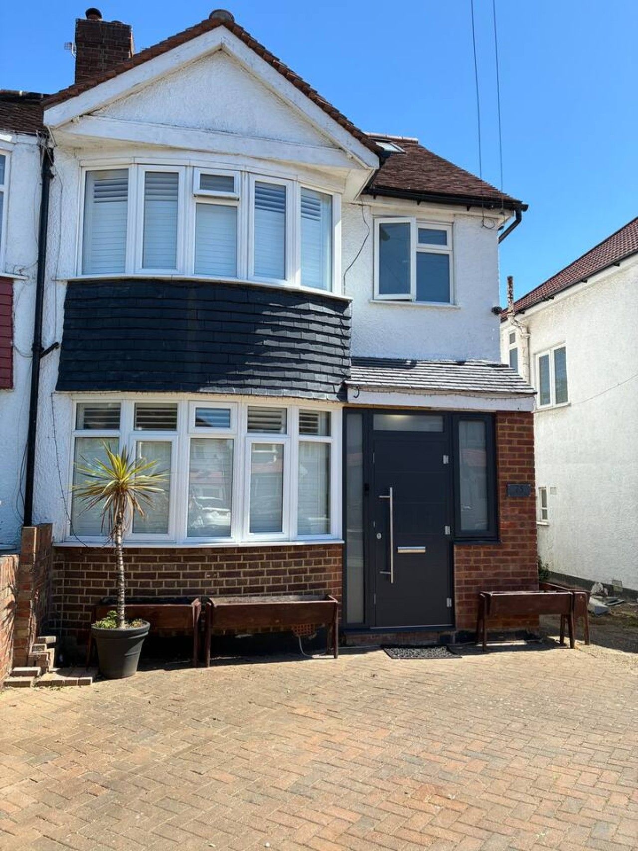 A two-story house with a black awning over the front door, a small garden with a palm plant, and a paved driveway.