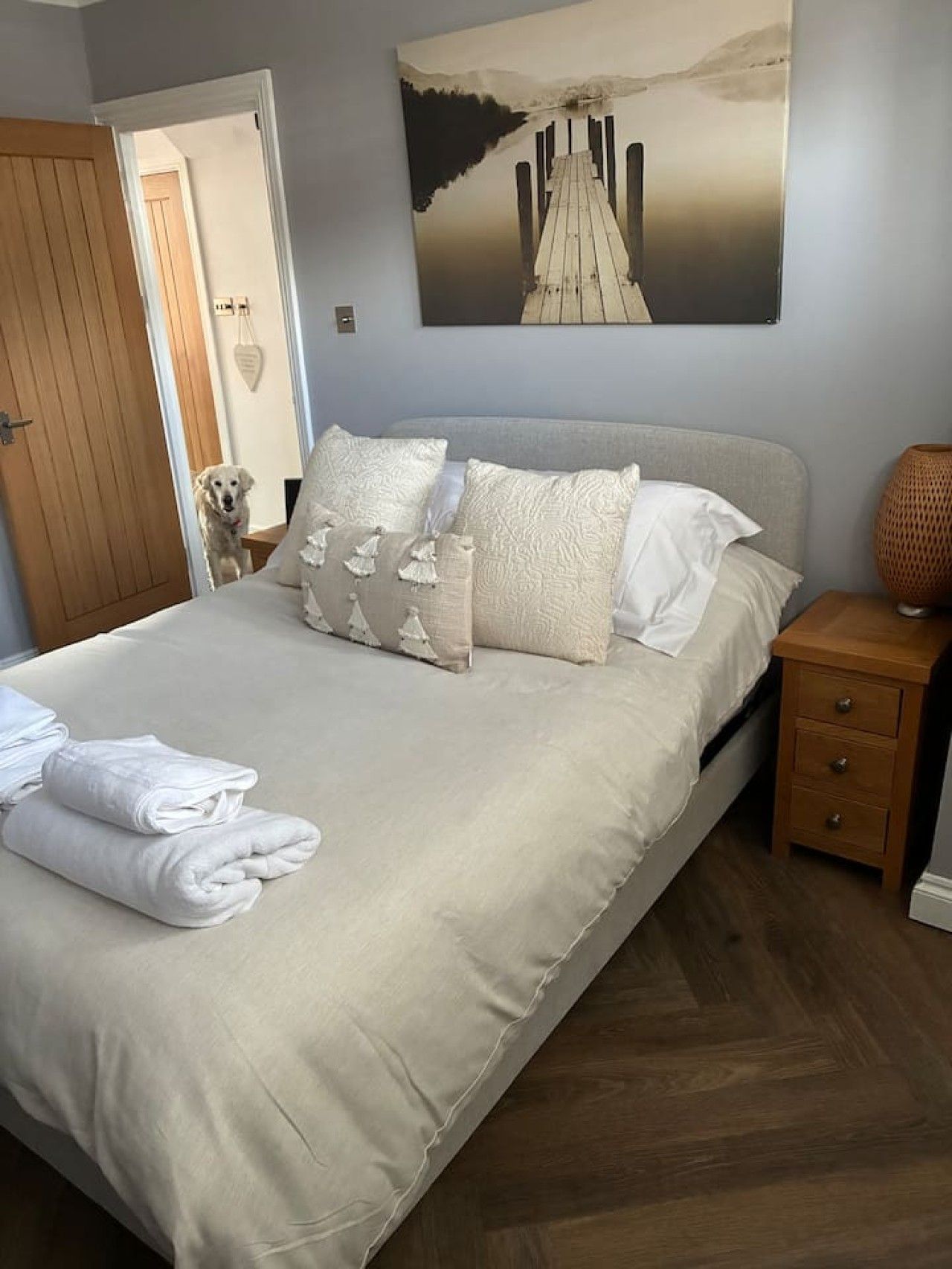 Ein ordentlich bezogenes Bett mit Kissen und gefalteten Handtüchern, ein Nachttisch aus Holz mit einer Lampe und ein Bild von einem Steg an der Wand.