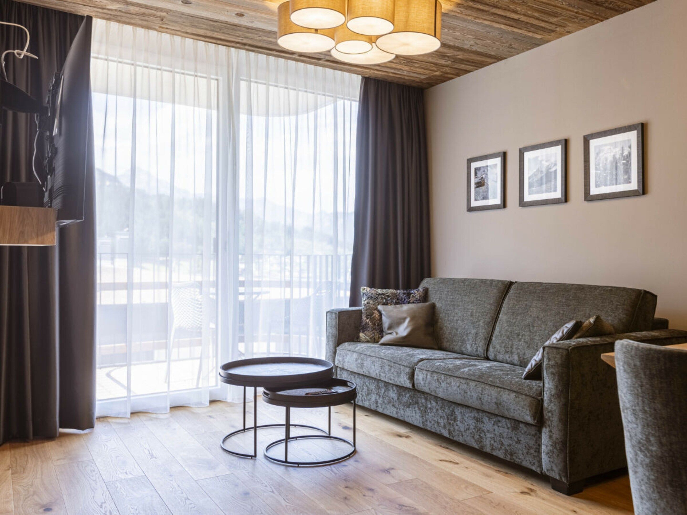 Uma sala de estar com sofá, duas mesas redondas, teto de madeira e uma janela grande com cortinas.