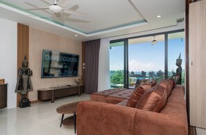 A modern living room with a large flat-screen TV, a sofa, a coffee table, and a ceiling fan. - Apartment With Sea View and Hot Tub on Balcony (Rawai)
