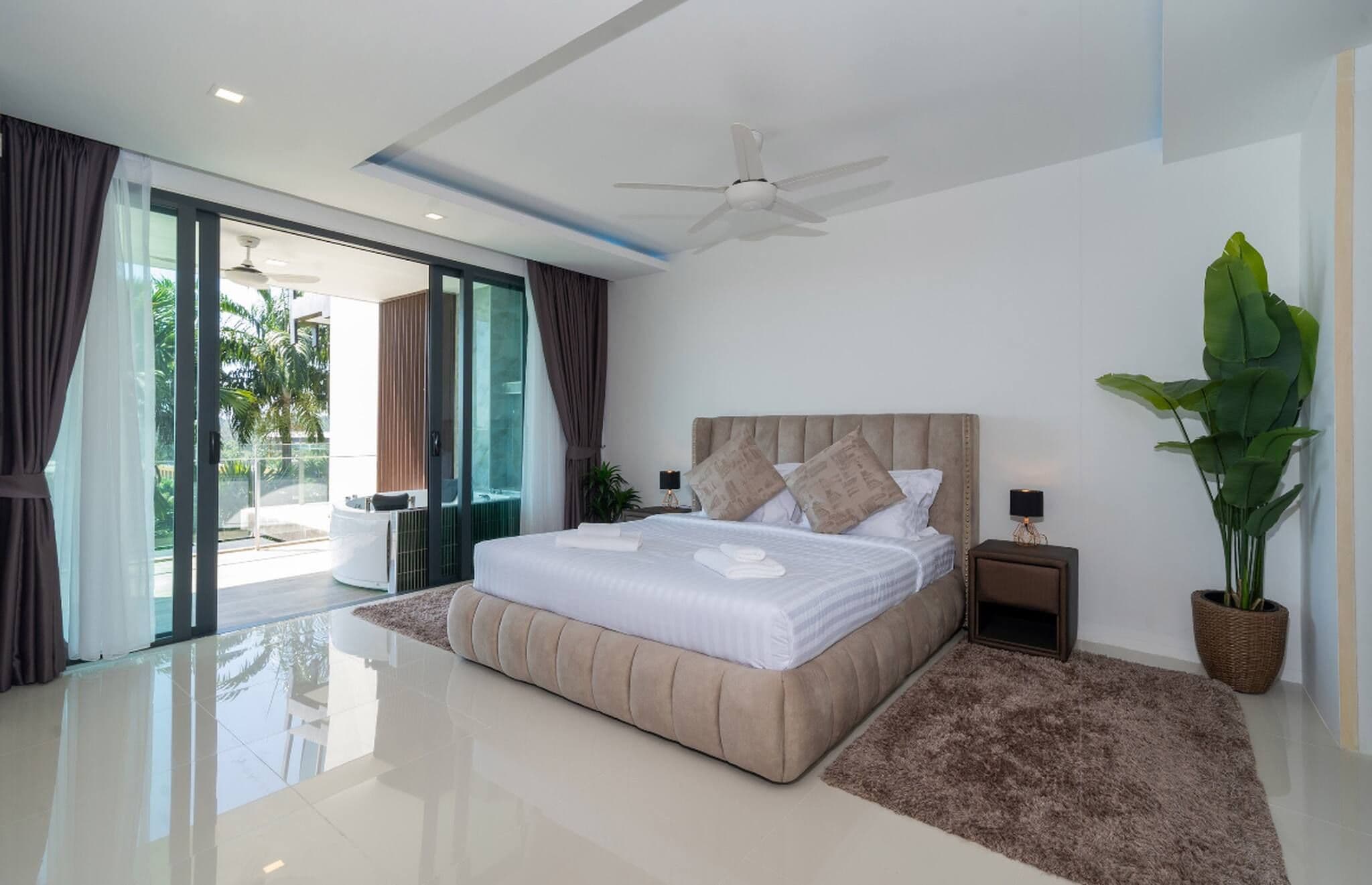A modern bedroom with a large bed, a ceiling fan, a potted plant, and a view of the outdoors.