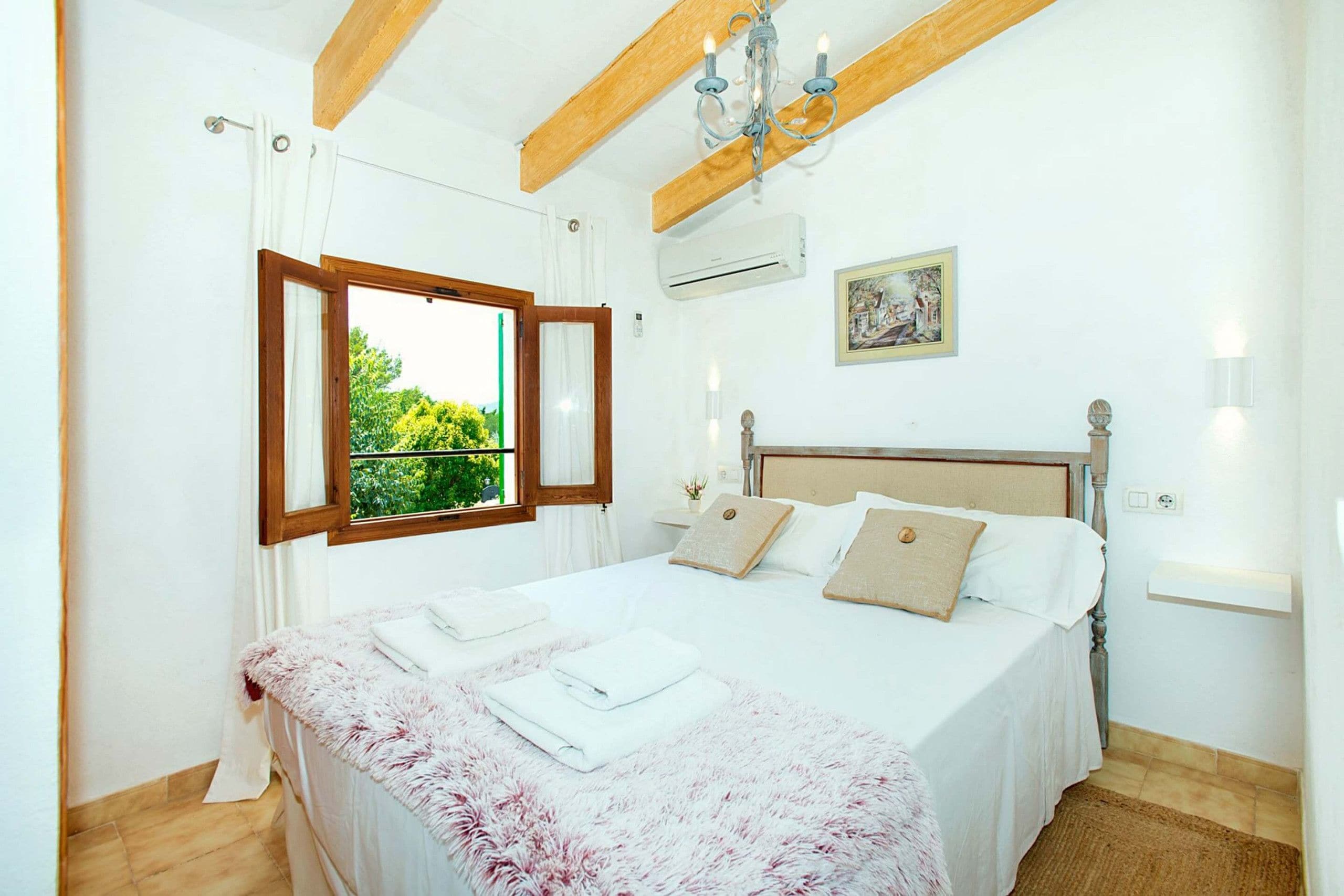 A bedroom with a bed, a window with curtains, a chandelier, and a painting on the wall.