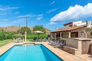 Una piscina amb gandules i una zona de terrassa, envoltada per un jardí i un turó.
