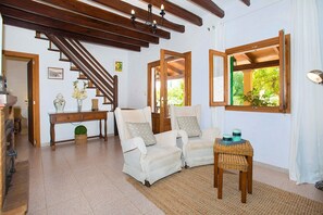 Ein Wohnzimmer mit einer Holztreppe, zwei weißen Sesseln, einem kleinen Tisch und einem Fenster mit Blick ins Grüne.