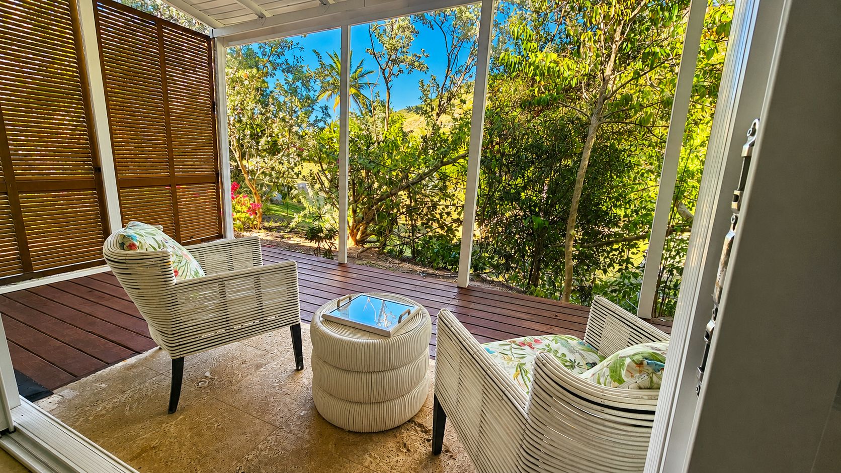 Un espacio de estar techado al aire libre con mobiliario de mimbre y una mesa con superficie de vidrio, con vistas a un jardín con árboles y arbustos.