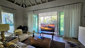 A living room with a television, a sofa, a coffee table, and a view of a landscape through the windows. - Private 2 Bed, 2 Bath Tropical Bungalow in upscale Christophe Harbour w Tennis (Saint George Basseterre Parish)
