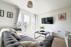 Ein Wohnzimmer mit einem grauen Sofa, einem Couchtisch, einem an der Wand befestigten Fernseher und einem Fenster mit Vorhängen.