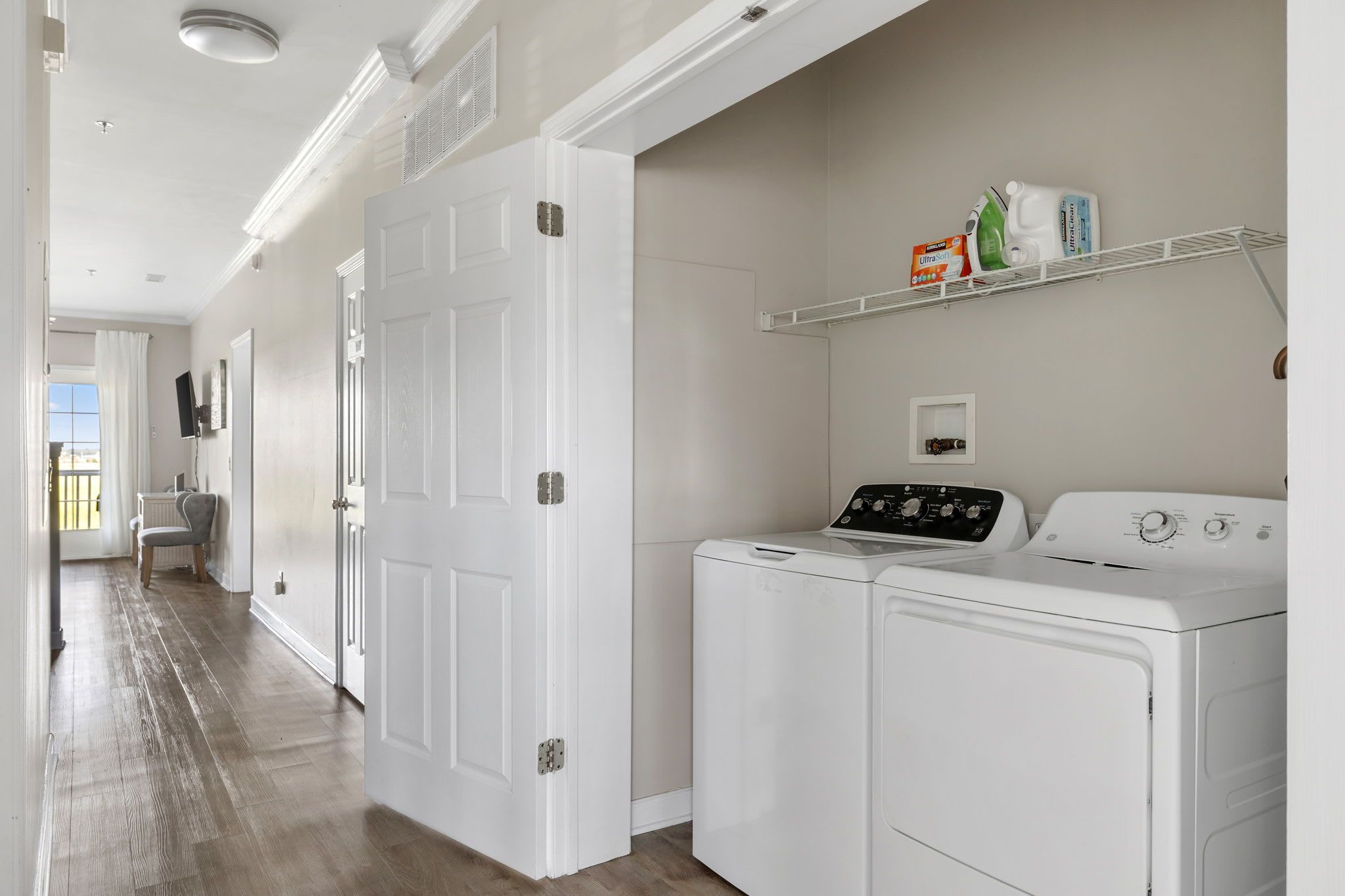 A laundry room with a washer and dryer, a shelf with laundry supplies, and a door leading to another room.