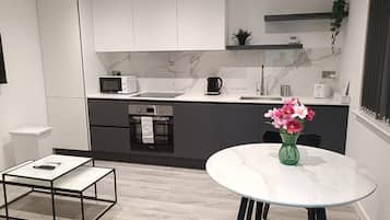 A modern kitchen with white cabinets, a microwave, a kettle, and a sink. There's a dining table with a vase of flowers and a coffee machine.