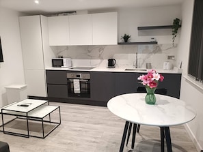 A modern kitchen with white cabinets, a microwave, a kettle, and a sink. There's a dining table with a vase of flowers and a coffee machine.