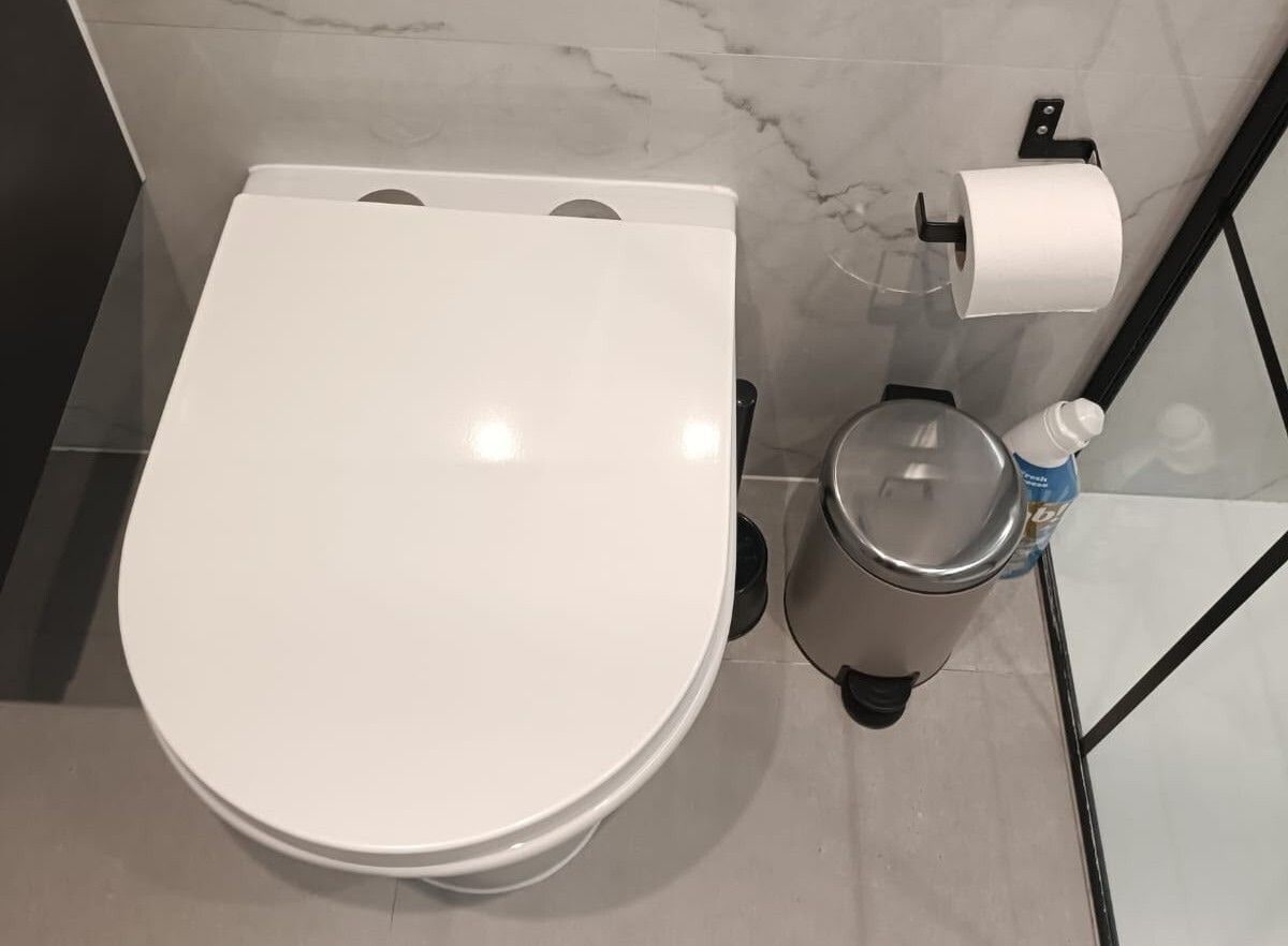 Des toilettes blanches avec un abattant, un porte-rouleau de papier toilette et une poubelle, dans une salle de bain aux murs en marbre.