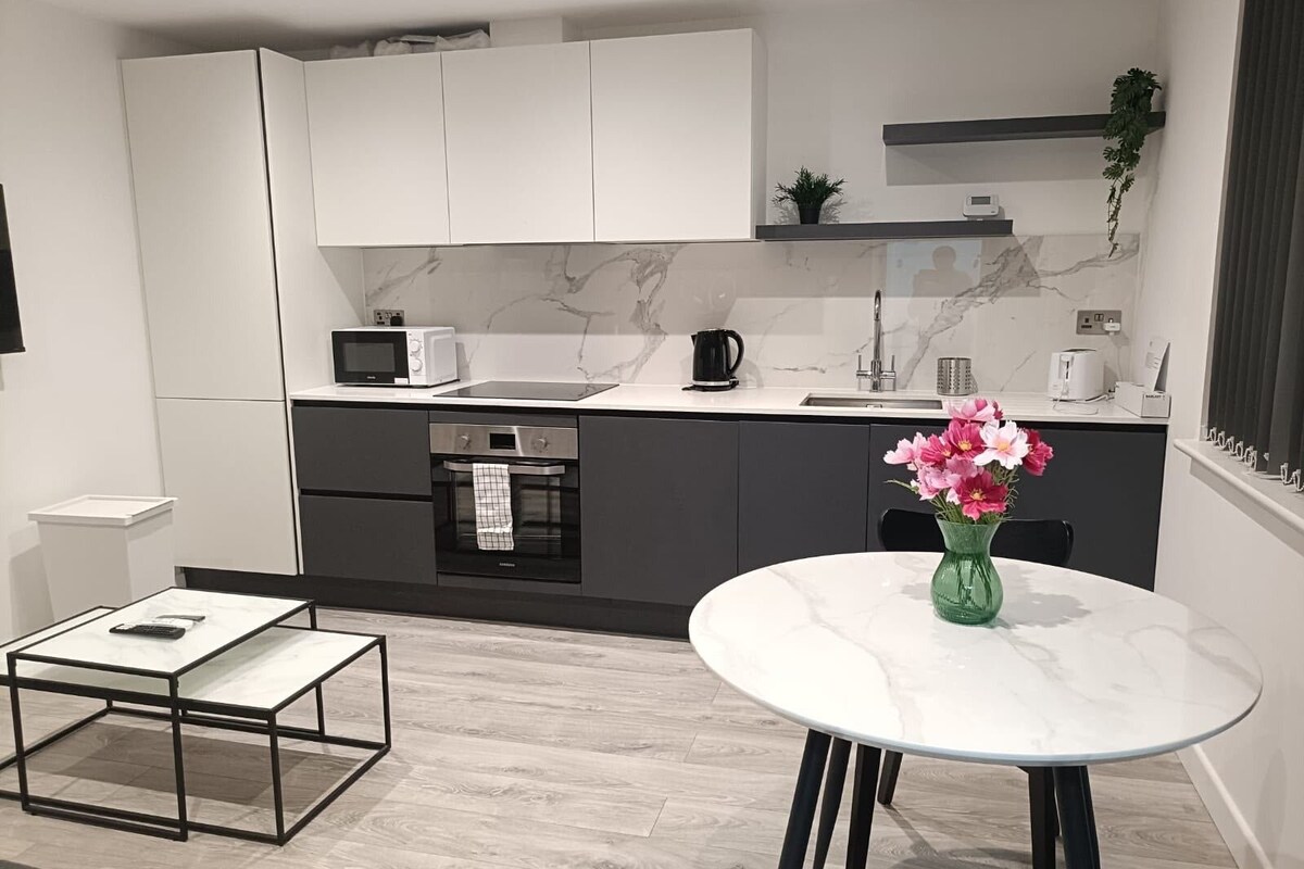 A modern kitchen with white cabinets, a microwave, a kettle, and a coffee maker. There's a dining table with a vase of flowers.