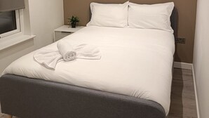 Een netjes opgemaakt bed met een grijze hoofdbord, witte beddengoed en een opgerolde handdoek op het bed.