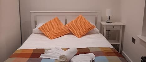 A neatly made bed with a colorful quilt, two orange pillows, and a rolled white towel.