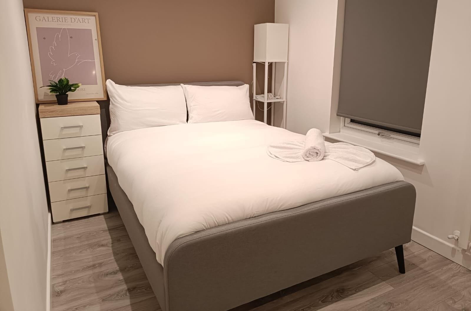 A neatly made bed with a rolled towel, a bedside table with drawers, a lamp, and a framed artwork on the wall.
